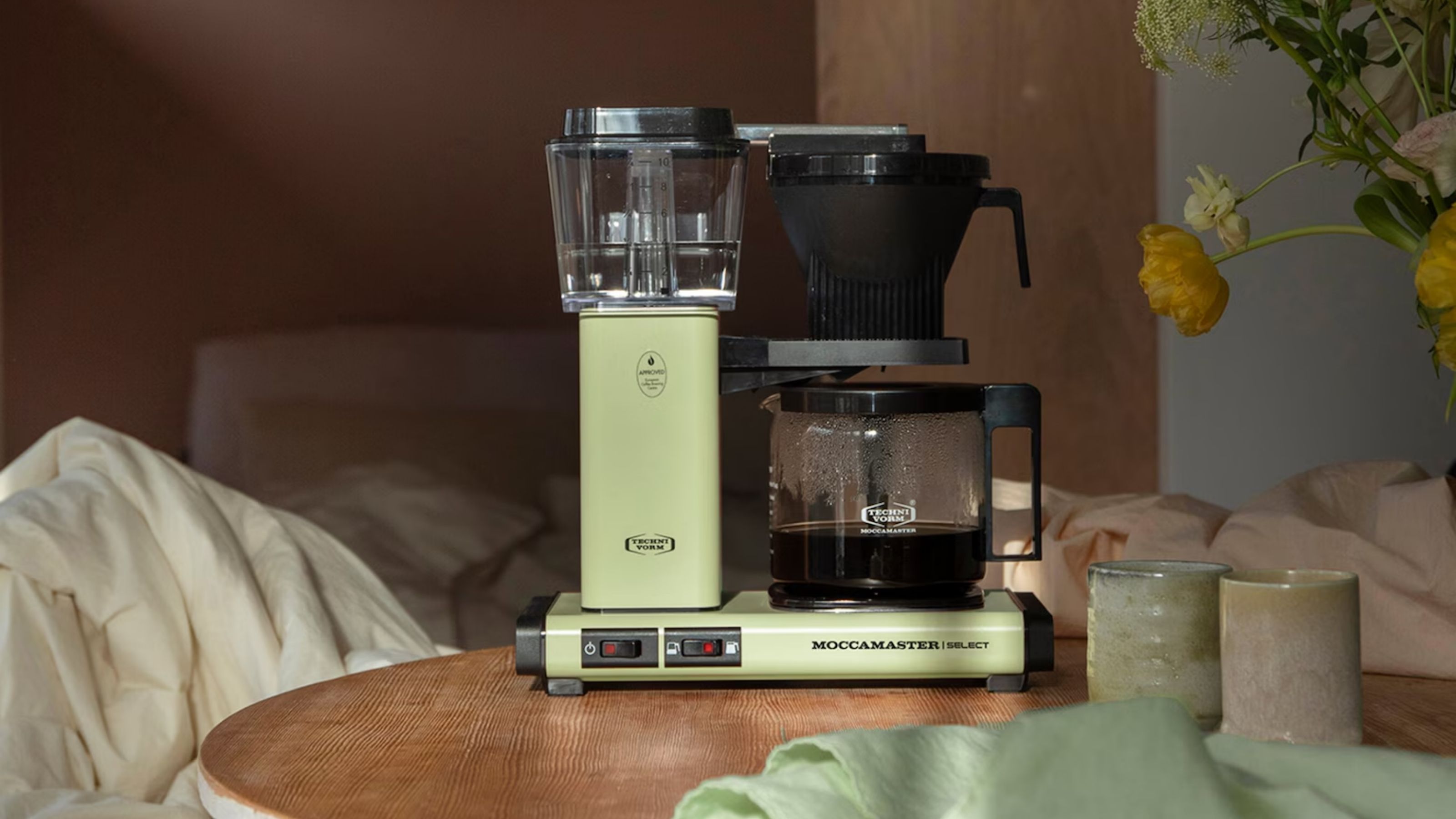Image of a pastel green Moccamaster drip coffee maker on a round wooden table in front of a messy, unmade bed.