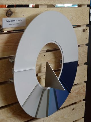 Sundial, by Patrick Nadeau for French design house Laorus. A white sundial with sections in shades of blue hanging on a wooden wall.