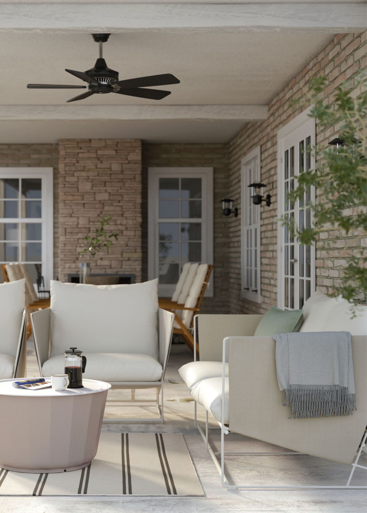 A garden seating area with a 2-seater beige sofa, two beige armchairs and a circular beige coffee table above a striped beige rug below a black ceiling fan and within a covered area of a brick home
