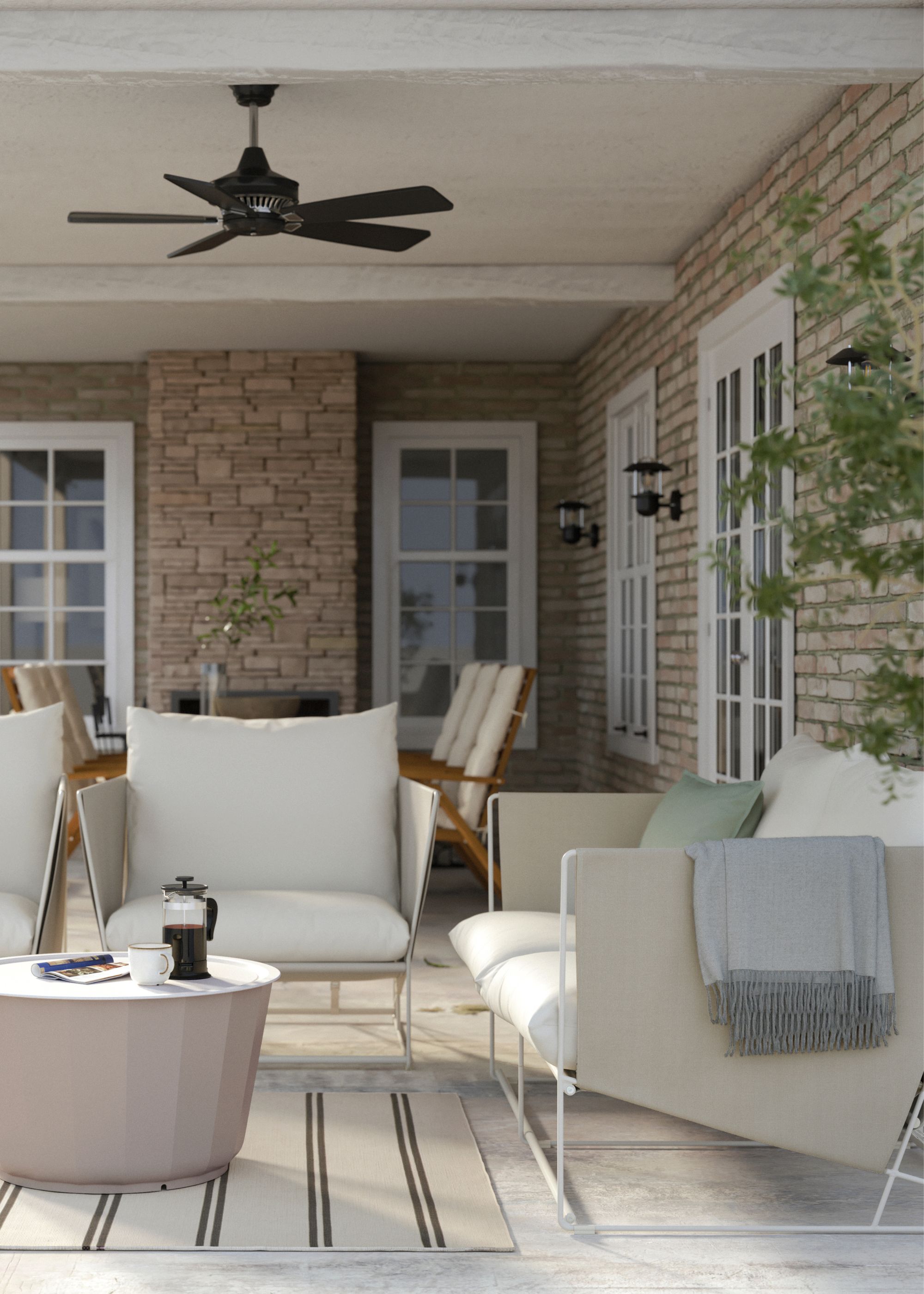 A garden seating area with a 2-seater beige sofa, two beige armchairs and a circular beige coffee table above a striped beige rug below a black ceiling fan and within a covered area of a brick home