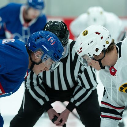 heated rivalry still of hudson williams as shane and connor storrie as ilya in their hockey gear on the ice ready to face off by a referee 