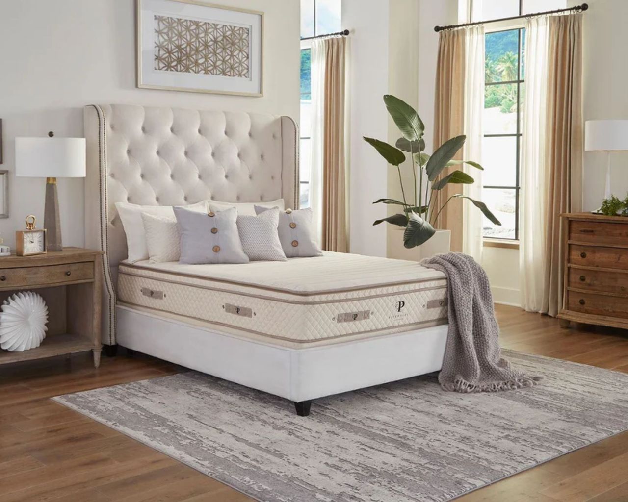 A PlushBeds Botanical Bliss Mattress is shown in a neutral on a cream bed with a large buttoned upholstered headboard, with neutral cushions on top. The floor of the bedroom is wooden, with a large grey rug beneath the bed. To the left of the bed is a wooden bedside table with a wooden lamp with a white shade, and to the left is a large crittal window with layered sheer and beige curtains, and a large houseplant, with a wooden chest of drawers. Above the bed is a large framed artwork.