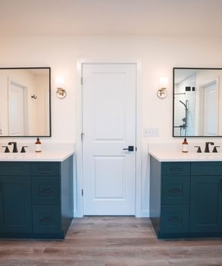 A cream colored bathroom with a white door, two square mirrors, and two identical sink units on the left and right hand sides with black faucets, brown soap, white surfaces, and dark blue bases