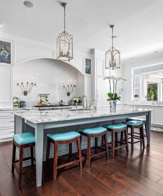 A large marble kitchen island with five bright blue stools in front of it, two large gold pendant lights above it, and a countertop and two gold wall sconces behind it