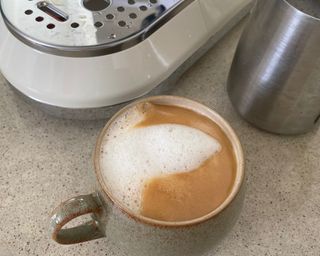 A cup of coffee alongside the Smeg coffee maker with metal milk jug