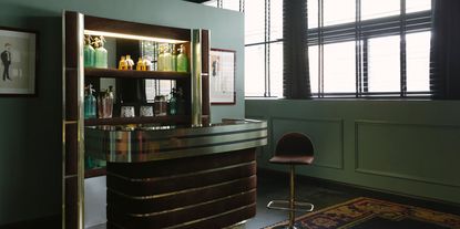 A sleek, brass and red velvet vintage bar features in a retro-fueled hotel room washed in mint green paint and decorated with framed artworks and rugs.