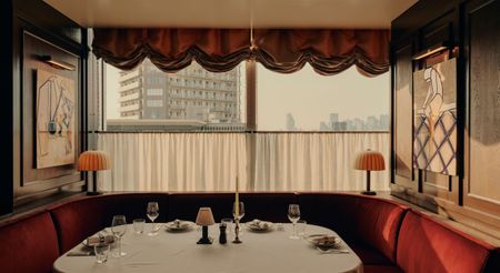 A restaurant table, decorated with red velvet banquettes, mushroom-shaped table lamps, and beautifully presented art, lets guests look outside a pair of cafe curtains and onto the city at dusk.