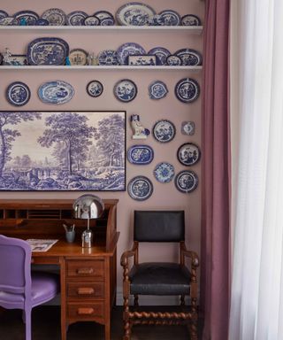 A light pink-purple room with an antique wooden desk, a purple chair, and a wall of antique porcelain plates