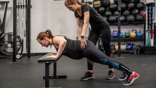 Woman performs incline press-up using a weights bench as a trainer guides her movement