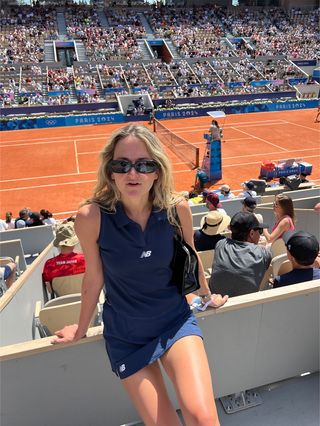 Eliza Huber wearing a navy blue New Balance tennis skirt set with sunglasses at Stade Roland Garros during a tennis match.