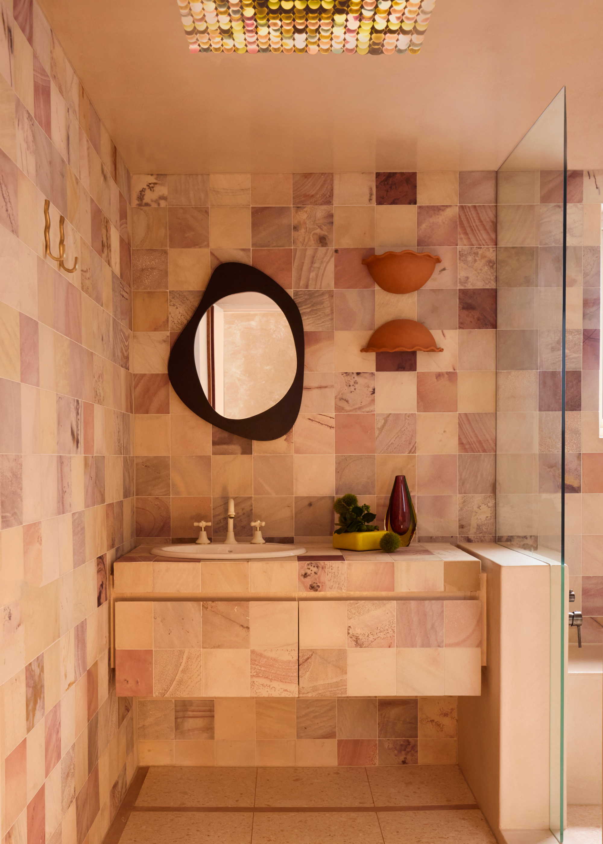 A modern bathroom with colorful pink-hued tiles, a floating vanity, and an abstract brown-framed wall mirror above with two terracotta-hued wall scones pointing in opposite directions (up and down)