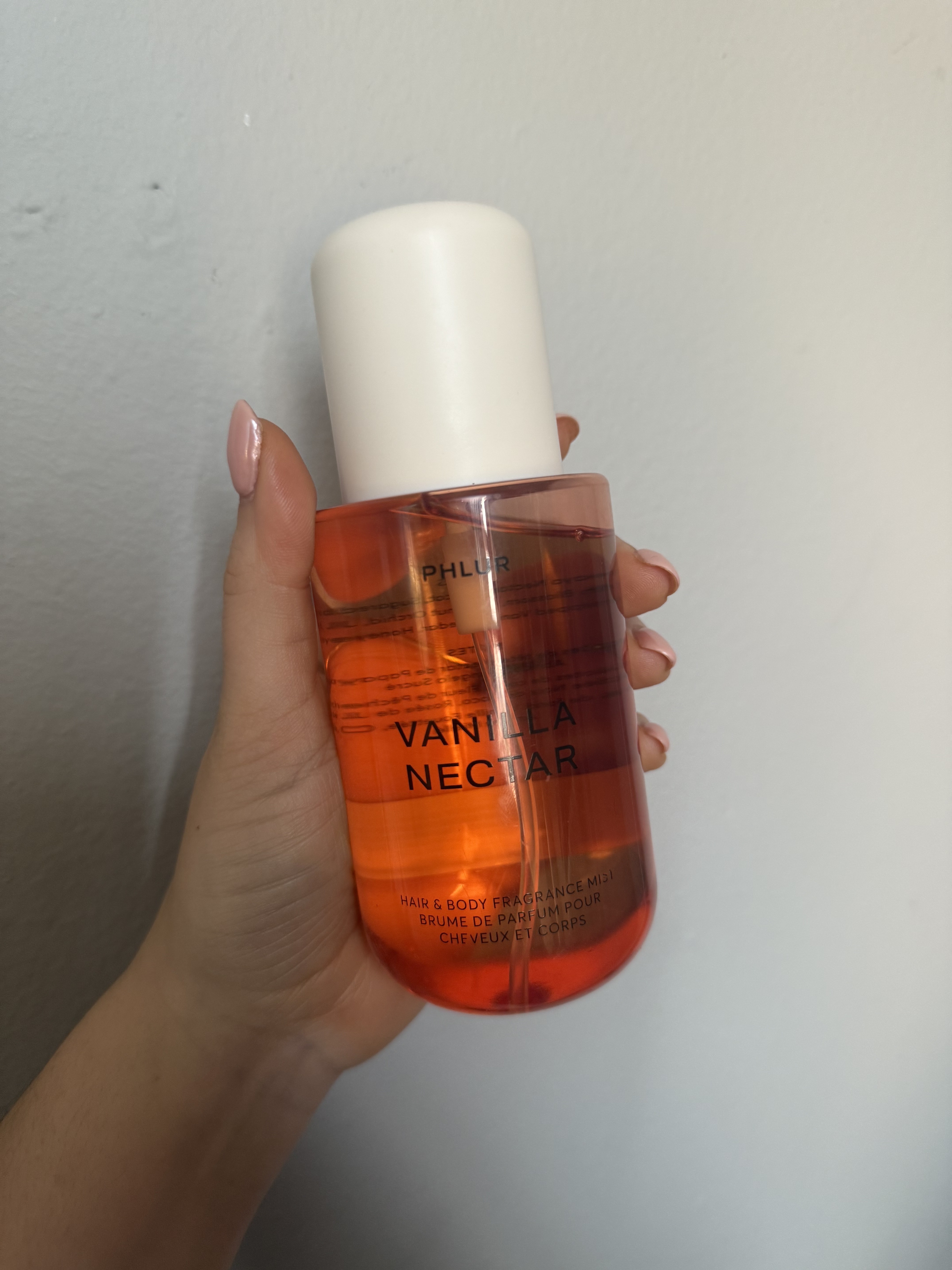 Brooke knappenberger holding up bottle of phlur vanilla nectar body mist against grey wall