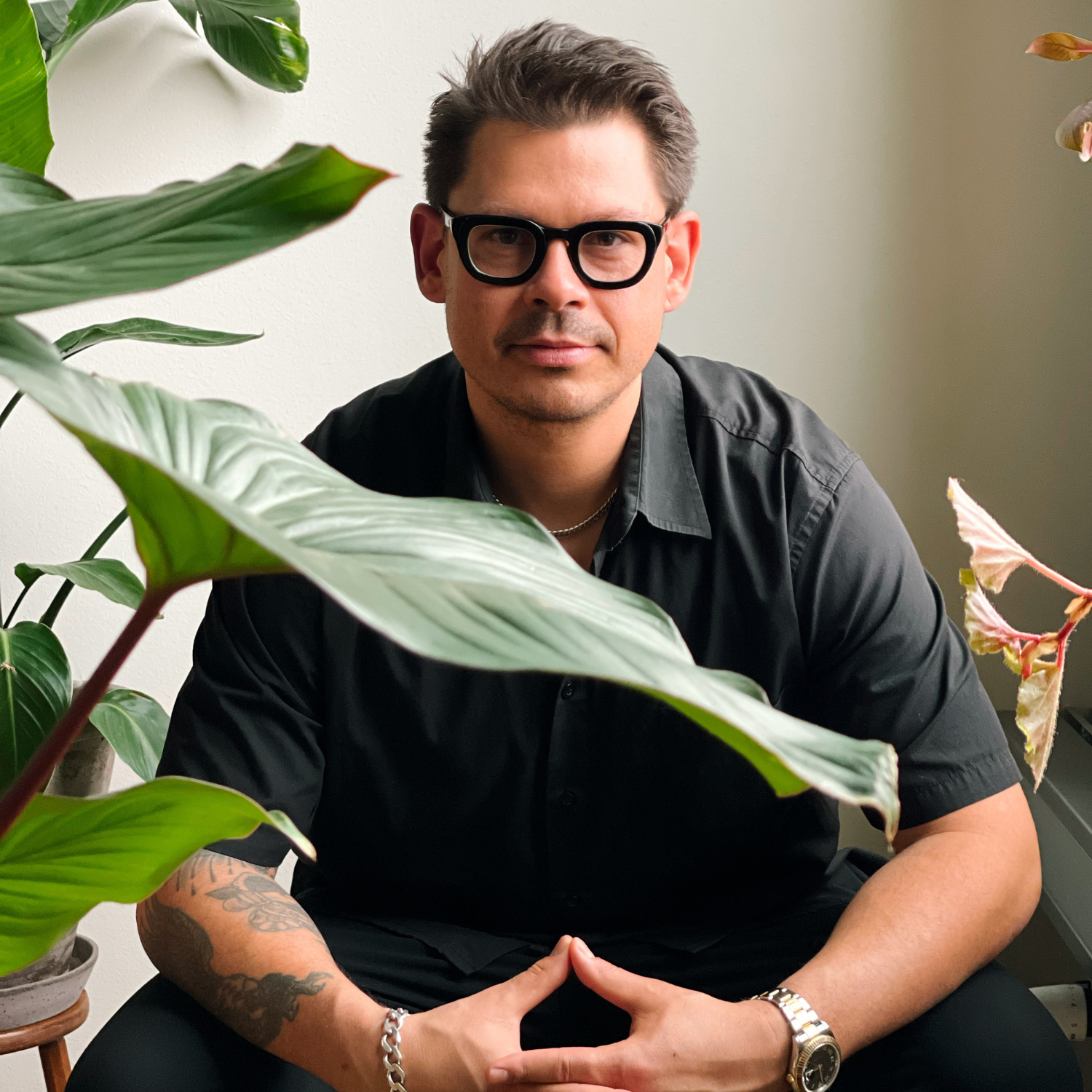A headshot of a man with short hair, thick black framed glasses, in a black shirt on a chair in front of leafy plants