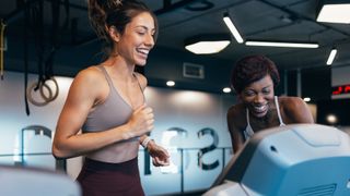 Two women laugh and smile as they run on treadmills.