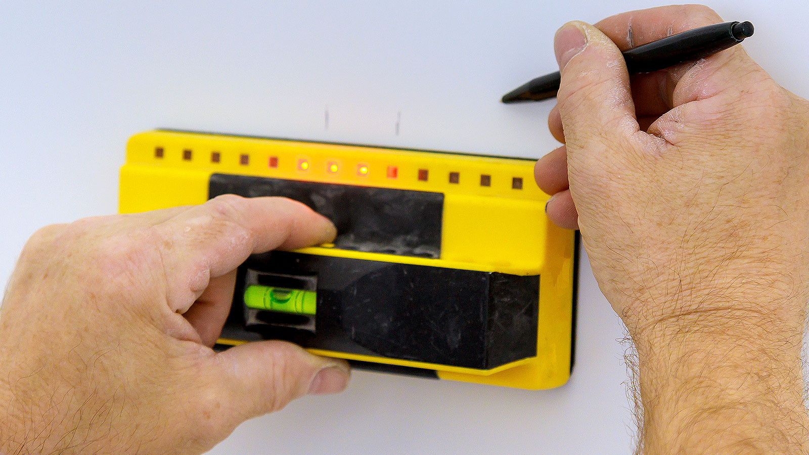 Pair of hands holding yellow electronic stud finder  against light wall and marking position of stud