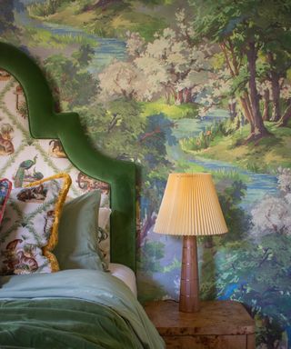 bedroom with green wood/forest style wallpaper mural with a curved headboard with green velvet trim, nightstand and lamp, green bedding