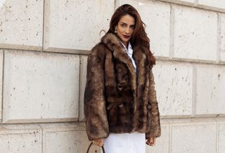 A woman wears a brown short fur coat, a white mini dress, and a brown top handle bag.