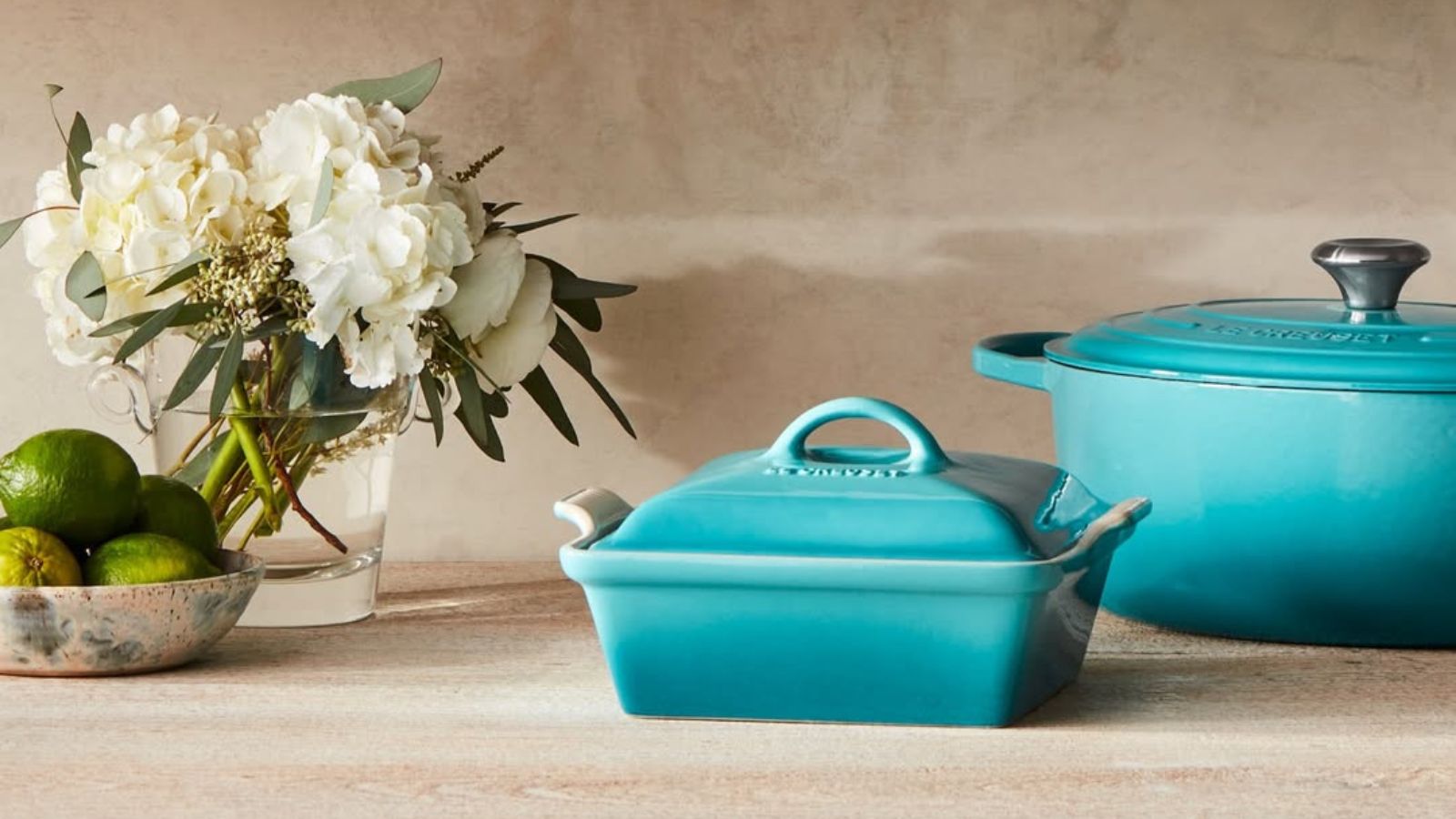 Le Creuset enamel Dutch oven and casserole dish in the Caribbean shade next to a vase of white flowers and a bowl of limes