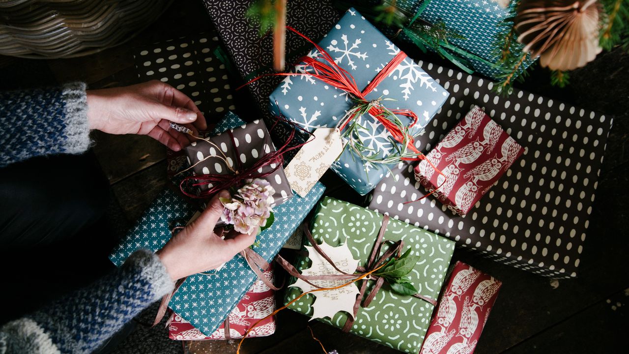 a selection of Christmas presents in various colours and with a variety of ribbon with a pair of hands adding the finishing touches