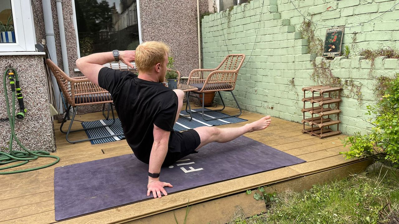 Fitness writer Harry Bullmore performs a kick through exercise in his garden on a yoga mat. He is concentrating, exercising low to the ground, with his body facing left. His right arm is straight behind him, with his right hand on the ground, while his left arm is bent and left hand close to his temple. His left leg is bent, and left foot resting on the floor, while his right leg is straight, pointing to the left, and elevated slightly off the ground.