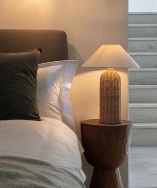 Rattan table lamp with cream cone shade, placed on sculptural stone side table next to bed