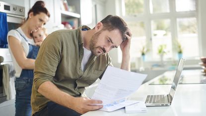 A man looks concerned as he looks at a letter at his kitchen counter while his wife holds their baby behind him.