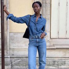 Influencer Sylvie Mus poses on a Paris sidewalk wearing a denim-on-denim look with a jeans button-down shirt, jeans, and a black shoulder bag