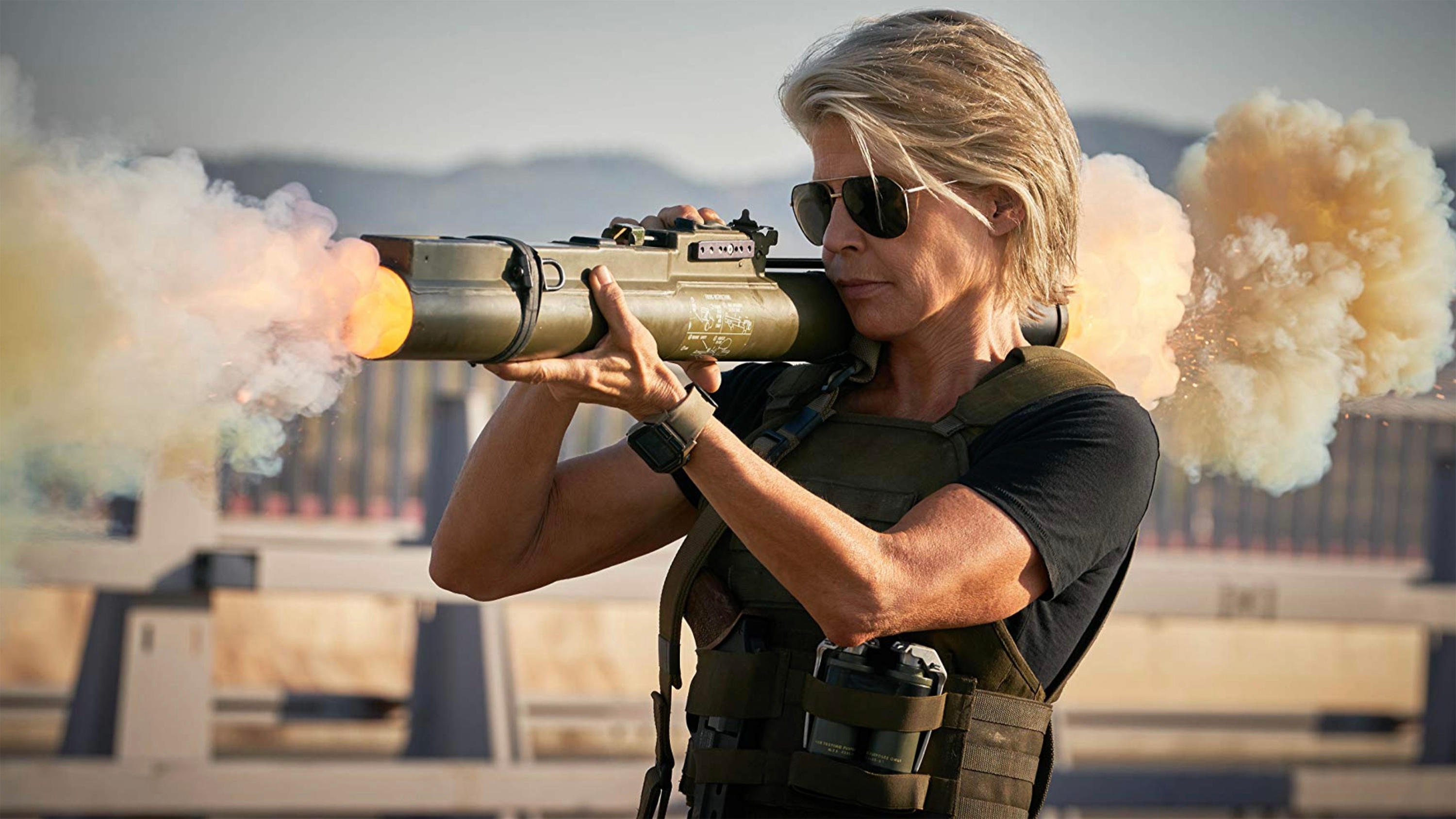 linda hamilton reprising her role as sarah connor as she holds a giant weapon on a military base covered in smoke in the 2019 movie terminator dark fate
