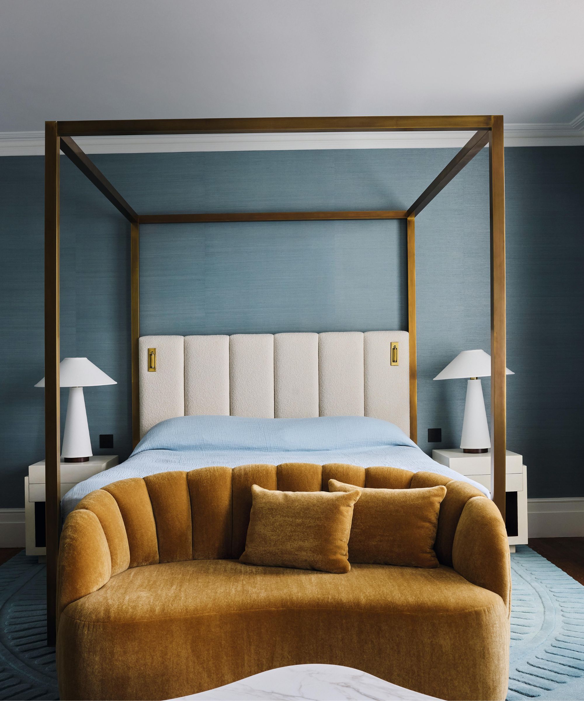 a blue grasscloth wallpapered primary bedroom with a four posted bed and mustard velvet sofa at the foot of the bed