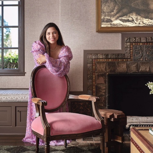 Image of a woman in a pink long-sleeve dress standing in front of a pink chair in a traditional looking interior.