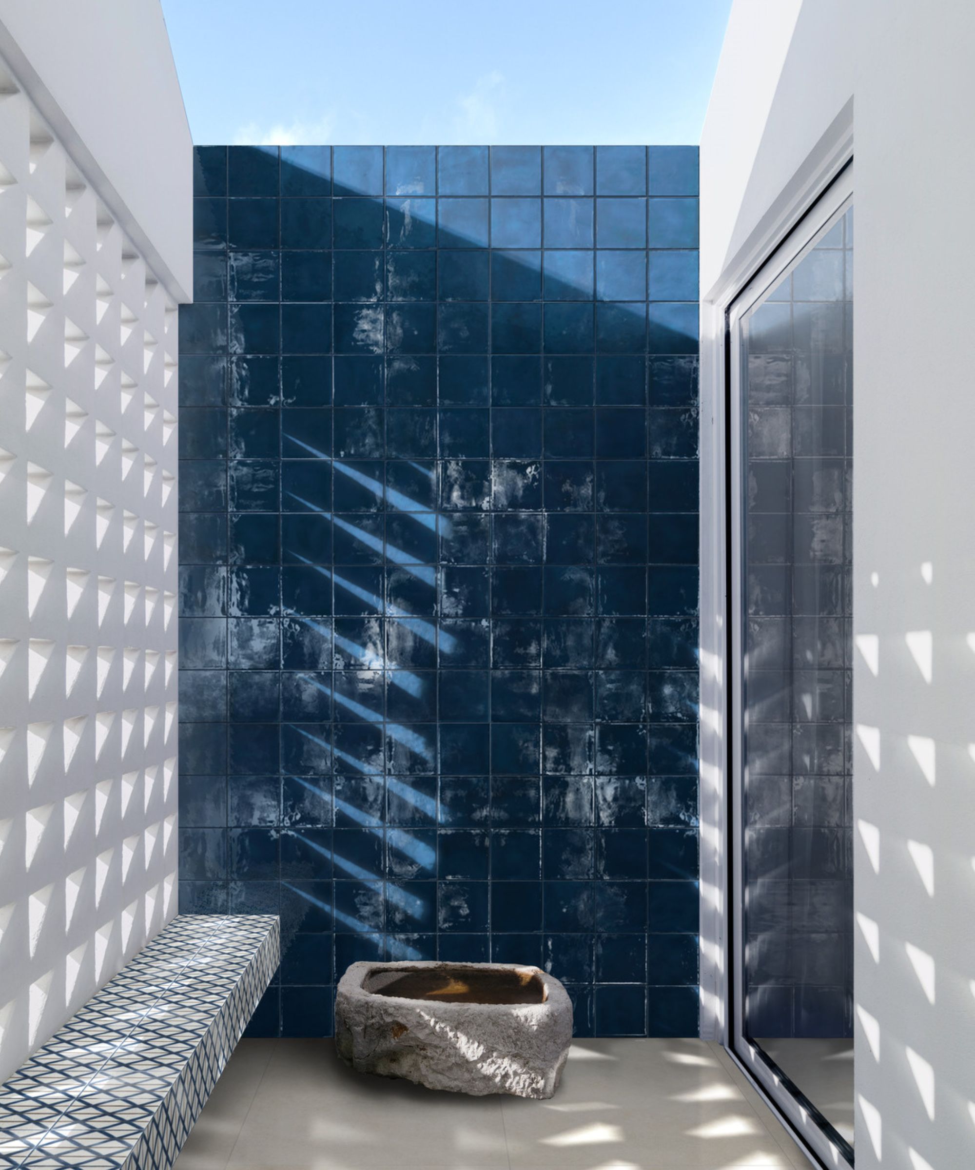 A narrow sun-drenched courtyard with a dramatic floor-to-ceiling deep navy zellige tiled feature wall, a carved stone trough at its base and a geometric patterned tiled bench