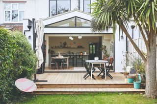 Lea-Wilson house: Rear of house with white extension, wood decking with steps down to grass, outdoor table and chairs