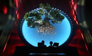 Close up view from below of the large round light display with an oversized bonsai tree hanging in front at The American Cathedral in Paris. There is a DJ booth, a DJ and another person beneath the display and the background is lit up red