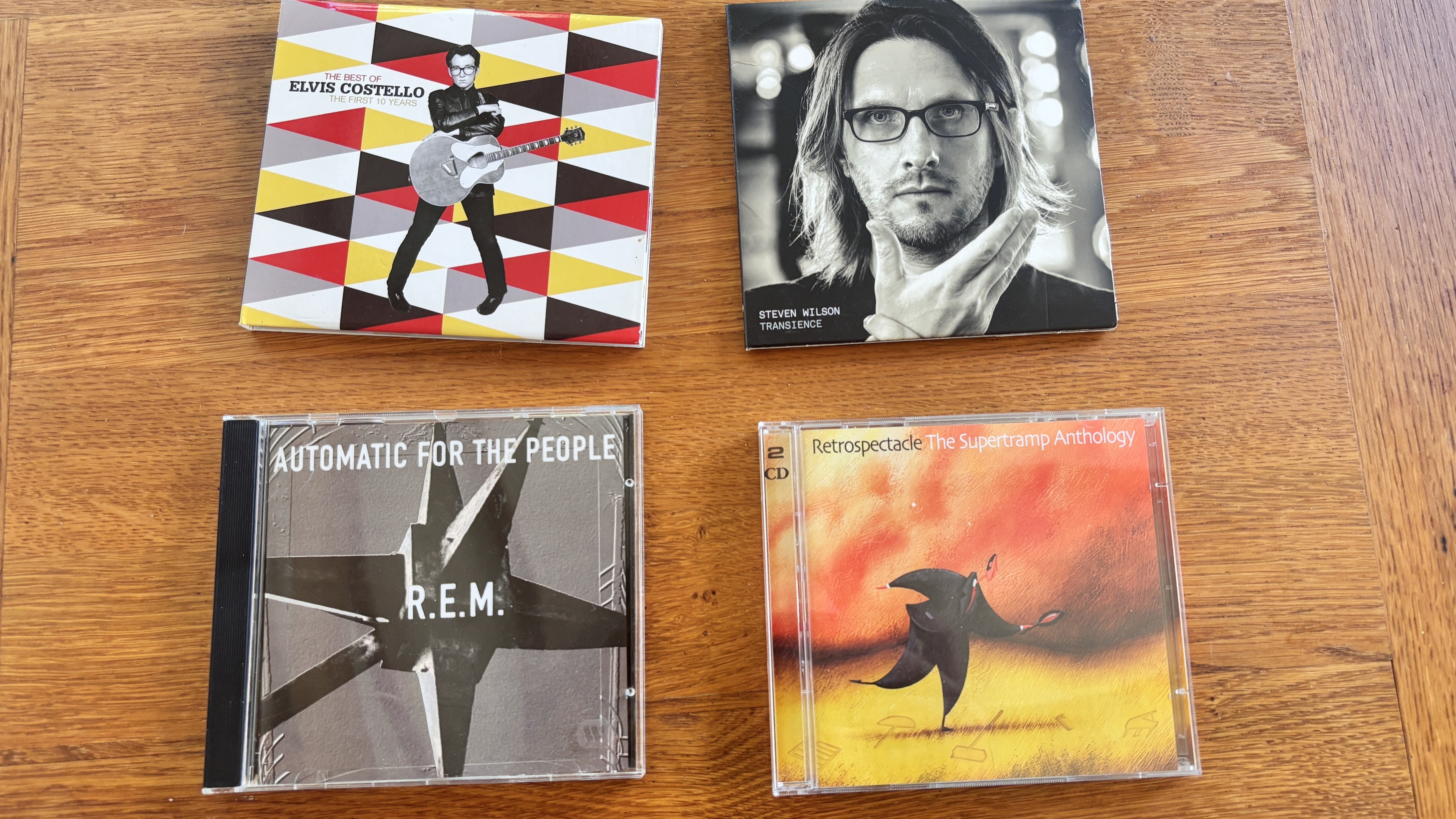 Four CDs on a wooden table, including REM, Elvis Costello and Supertramp