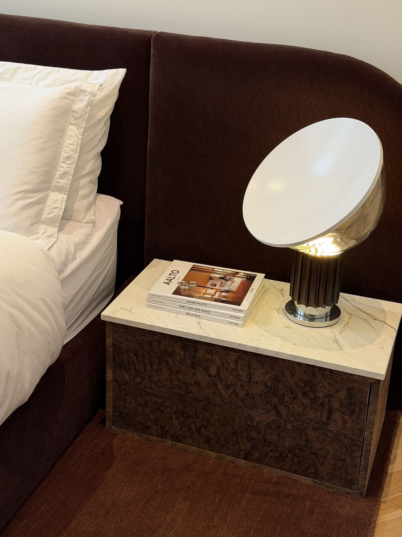 Image of a burl wood and white marble bedside table next to a bed with white bedding and a dark red, velvet extended headboard and matching rug.