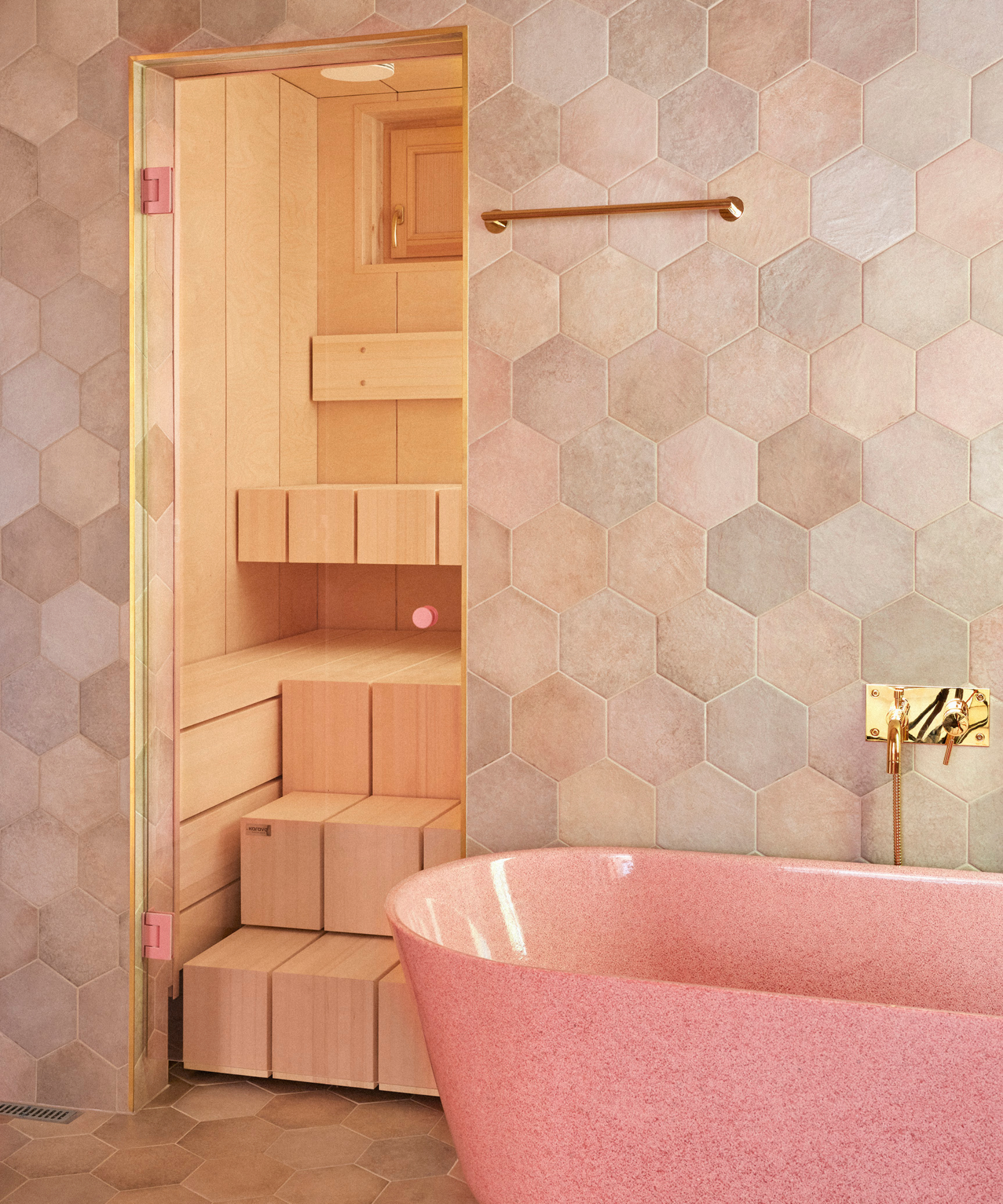 modern bathroom with hexagonal tile effect walls in shades of pink and grey, door leading to wooden sauna and pink freestanding bath with gold taps