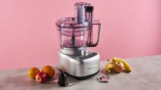 The Cuisinart Expert Prep Pro food processor pictured against a pink background. The small chopping blade is pictured to the left of the processor, and the spiralizing accessory is just in front.