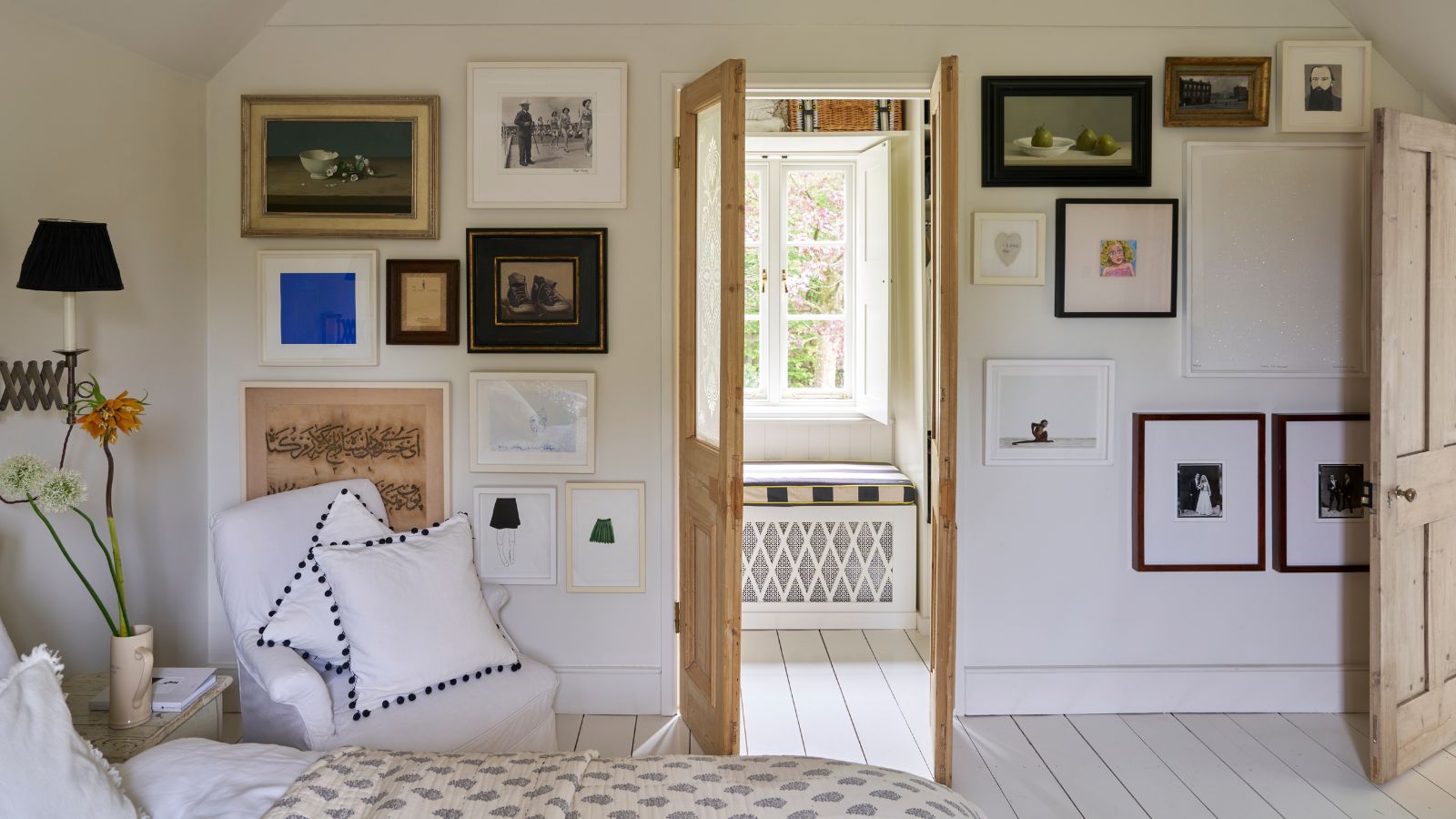 Bright bedroom with a gallery wall and bare wooden doors open to reveal a seated nook with window bench, cushion and white, crittal window