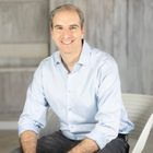 a photo of professional organizer Ben Soreff sat on a white chair against a wooden background, wearing a light blue collared long sleeve shirt and black jeans