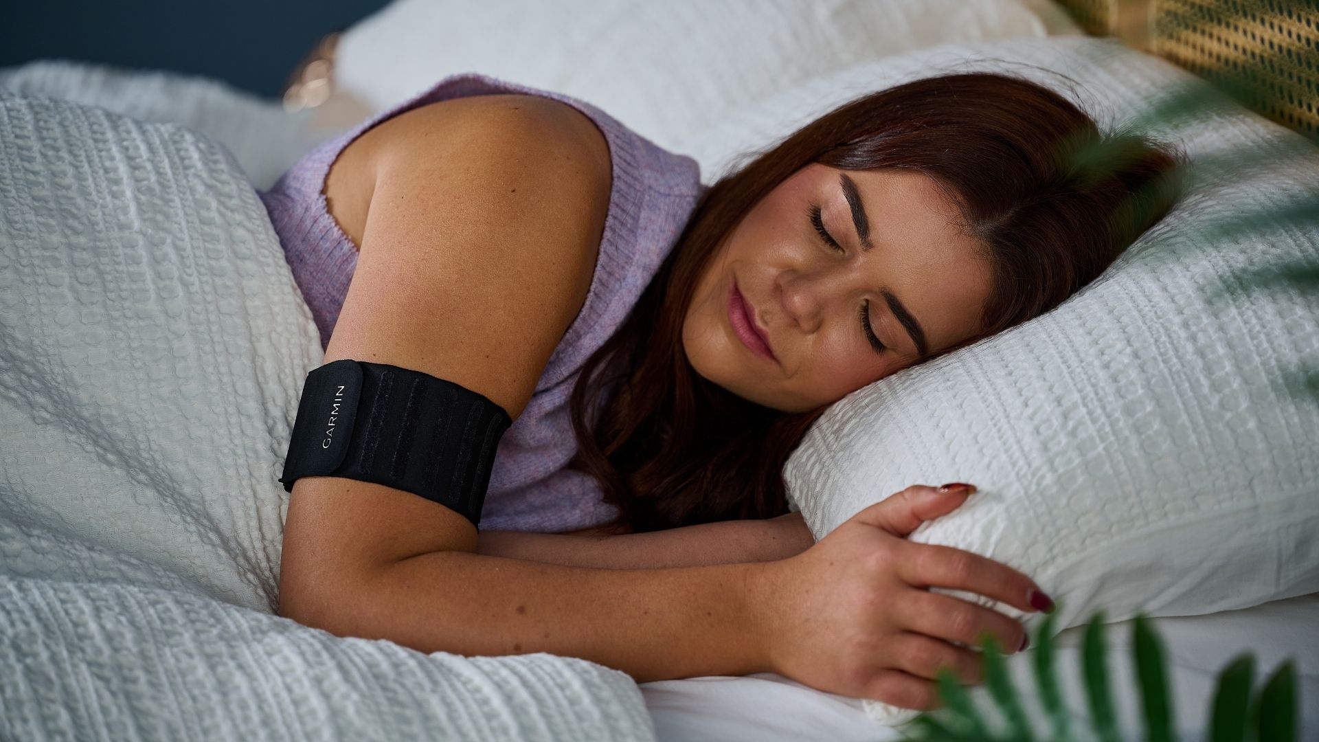 Our sleep tech reviewer laying in bed with white sheets wearing purple jumper and the black Garmin Index Sleep Monitor on right arm