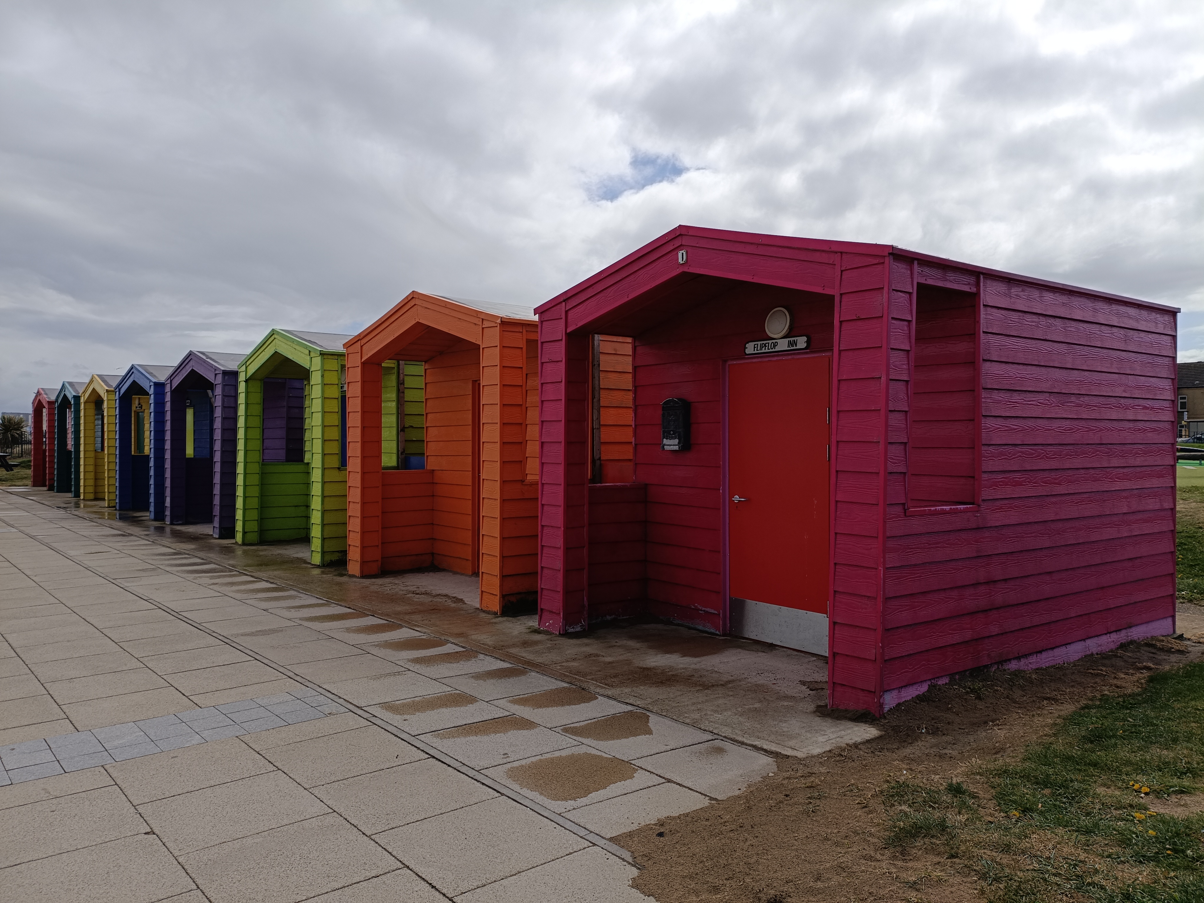 Row of brightly painted beach huts in various colours—pink, orange, green, purple, blue, and yellow—lined up along a paved promenade with grass and cloudy skies overhead
