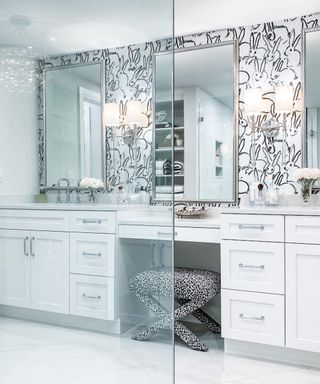 A bathroom with black and white patterned wallpaper, two silver rectangular mirrors, and a long white vanity with drawers and chrome handles with a leopard print stool underneath
