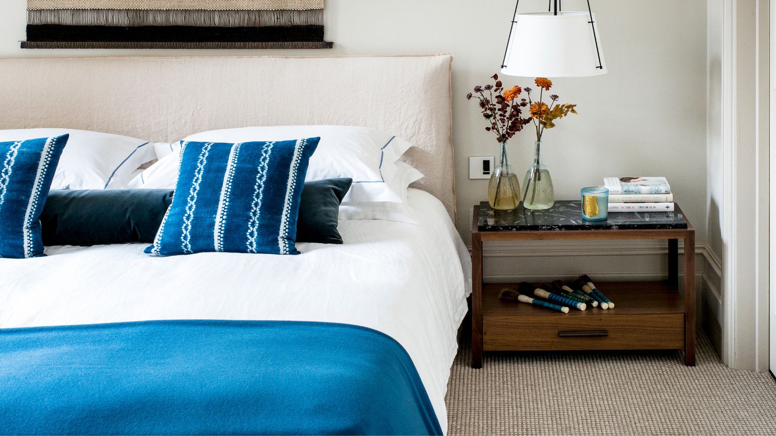 White bedroom with large double bed with blue bedding, a white lamp, dark wood nightstand, two glass vases filled with dried flowers, and a large wall tapestry. The carpet is light jute.