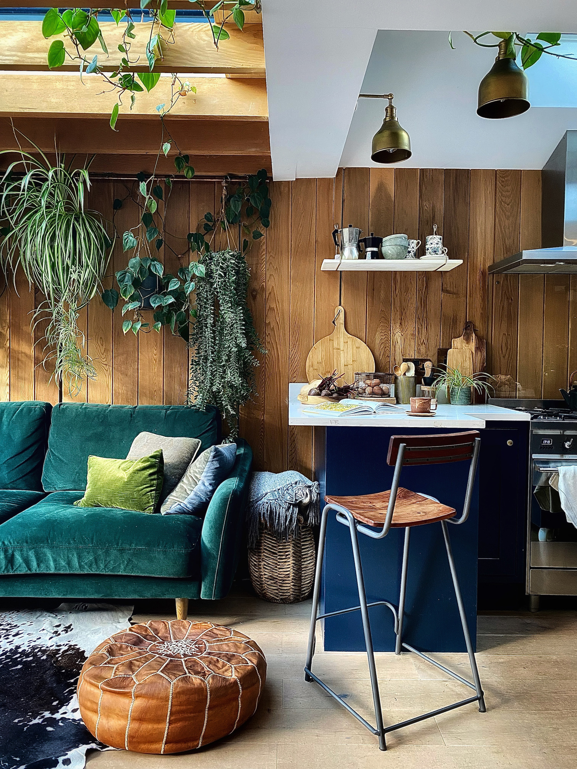 A kitchen/living space with wooden clad walls and multiple skylights. The kitchen cabinets are navy blue and the teal of the sofa is complemented by all of the hanging plants in the space.