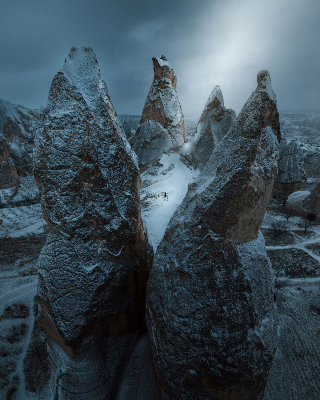 A dramatic winter landscape featuring towering rock formations covered in snow, with a lone figure standing amidst them