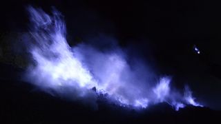 Night time image of blue flames produced by sulfurous gas at Kawah Ijen volcano in Indonesia. We see bluish-purple flames against a black background.