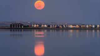 supermoon appearing orange/red above a row of houses. The moon is reflected in the water body below.
