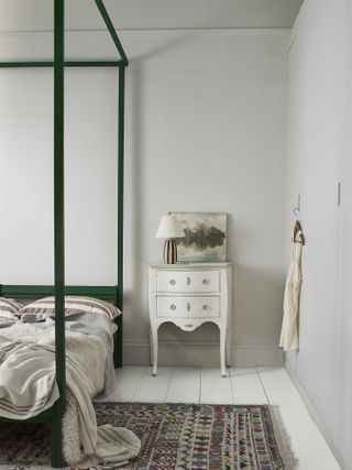 Bedroom with four poster, white painted floorboards and area rug, and white walls, green four poster bed, patterned rug, artwork, lamp