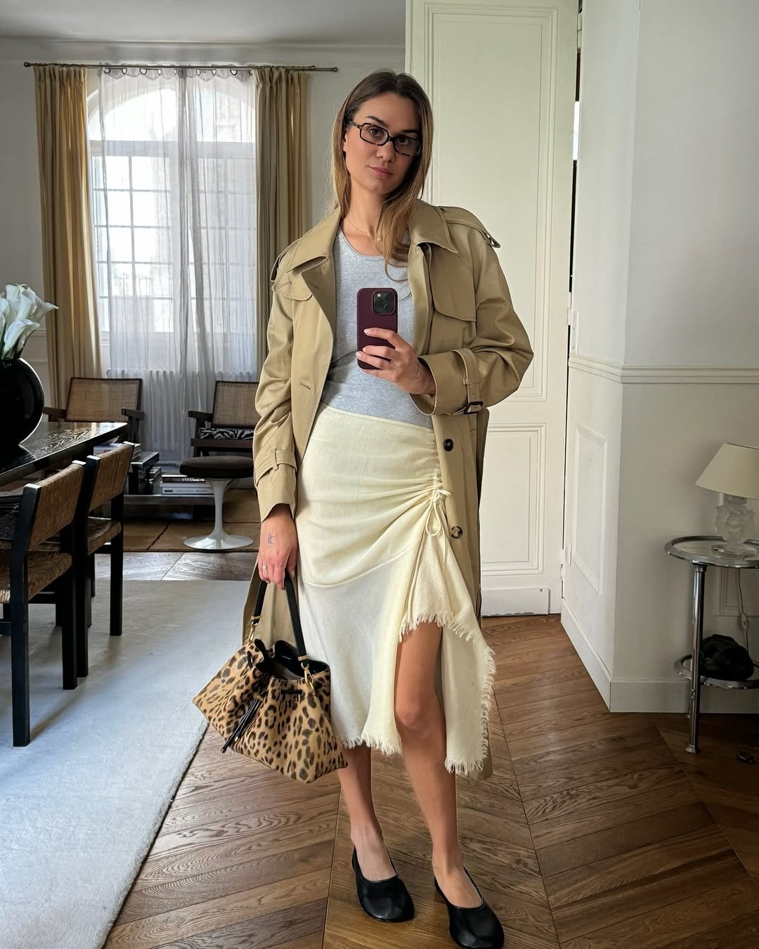 A woman standing in front of a mirror wearing a butter yellow asymmetrical skirt, a gray tee, and a trench coat.
