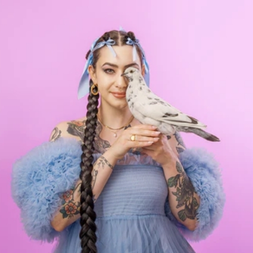 Image of a woman with tattoos wearing a light blue tulle dress with matching ribbons in her hair. Her hair is in a long braid and she is standing in front of a magenta background. She is holding a white pigeon up to one of her eyes.